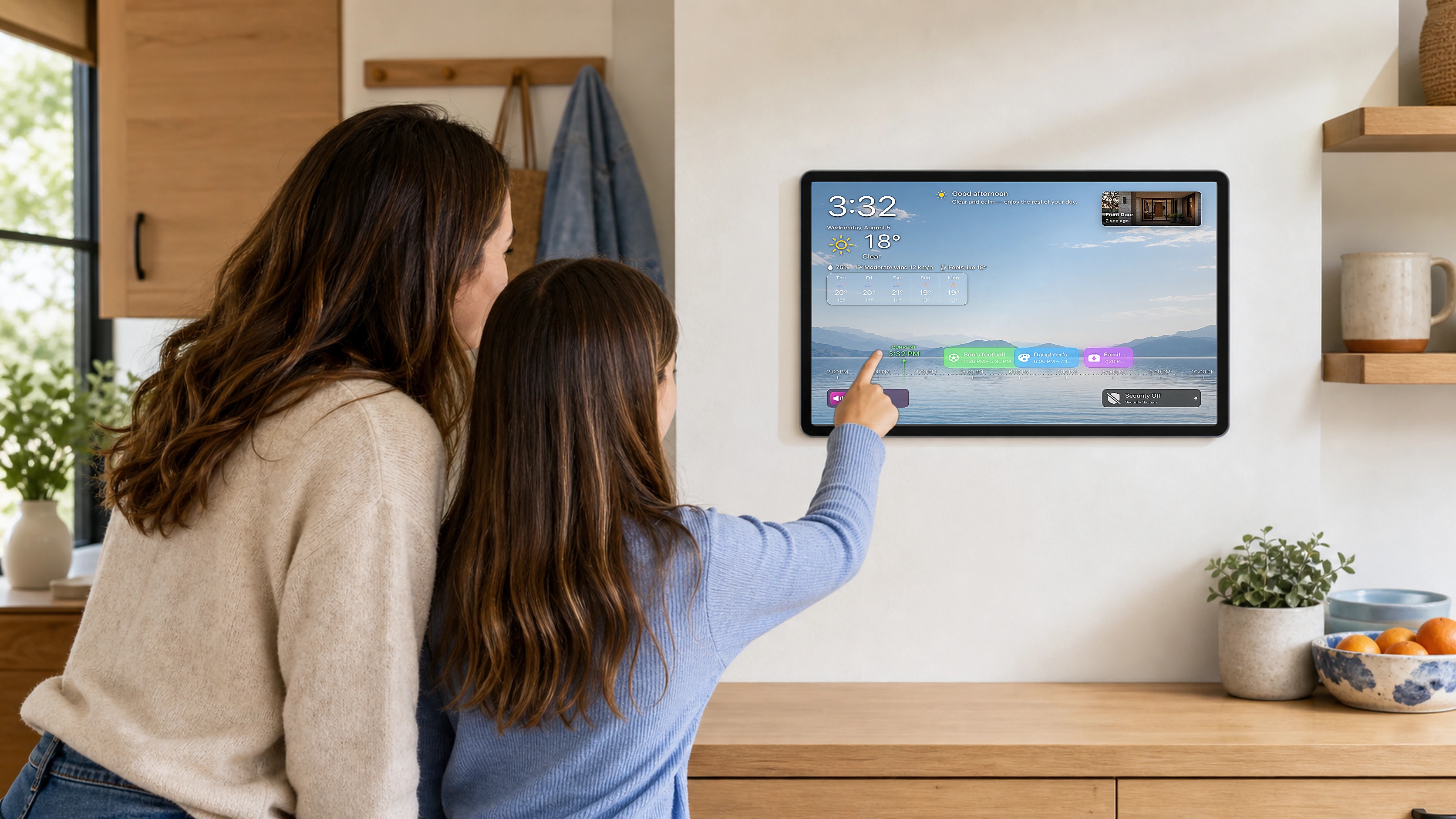 A mother and daughter checking their family calendar on a wall-mounted iPad in a sunny kitchen